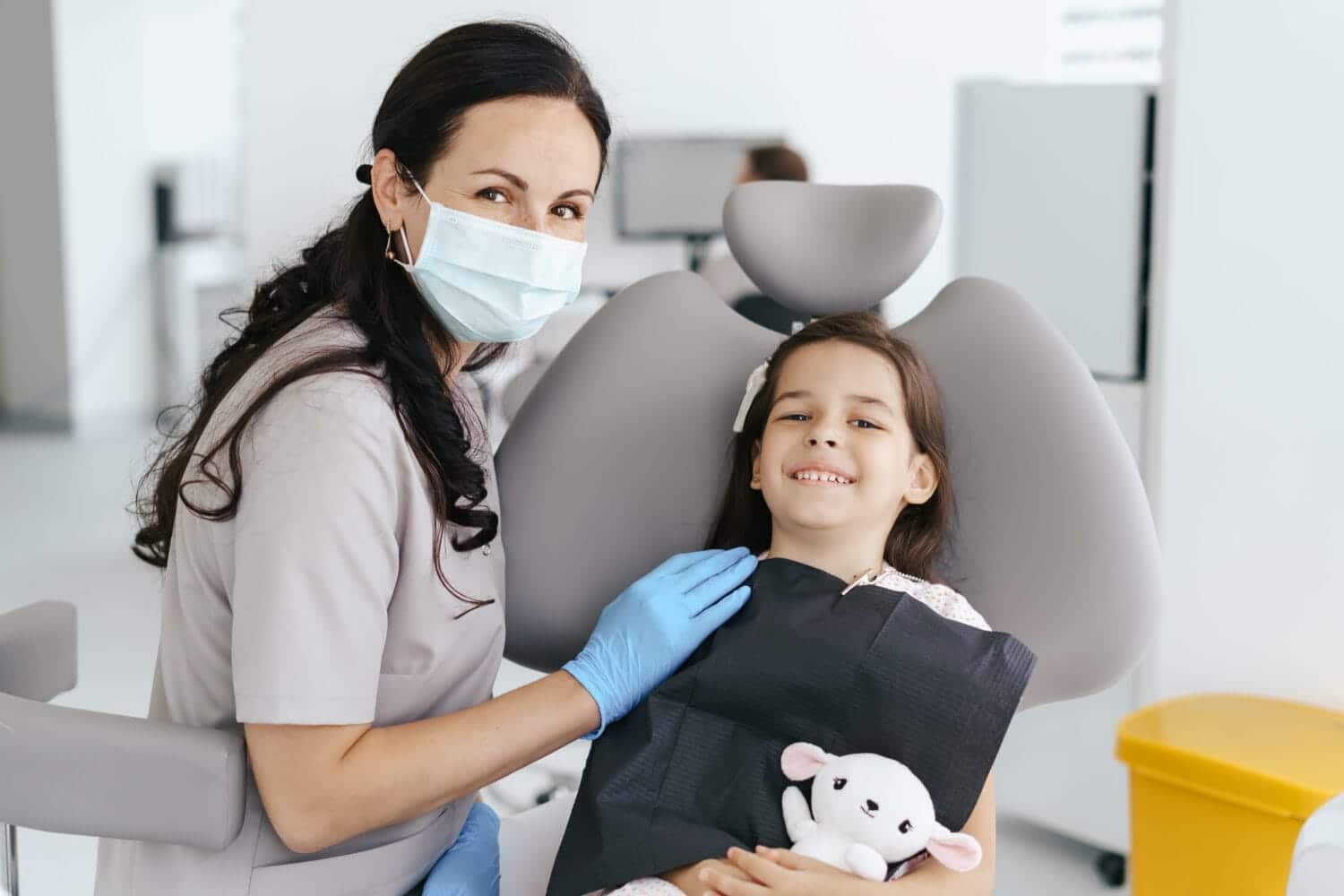 Pediatric dentist wearing a mask and gloves treating a smiling child patient in a dental chair during a comfortable visit.