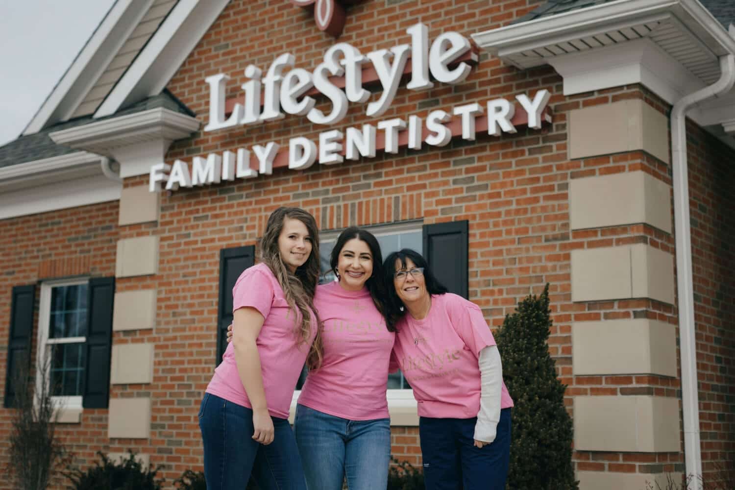 Dental team wearing matching shirts standing outside a family dentistry office, smiling in front of the building entrance.